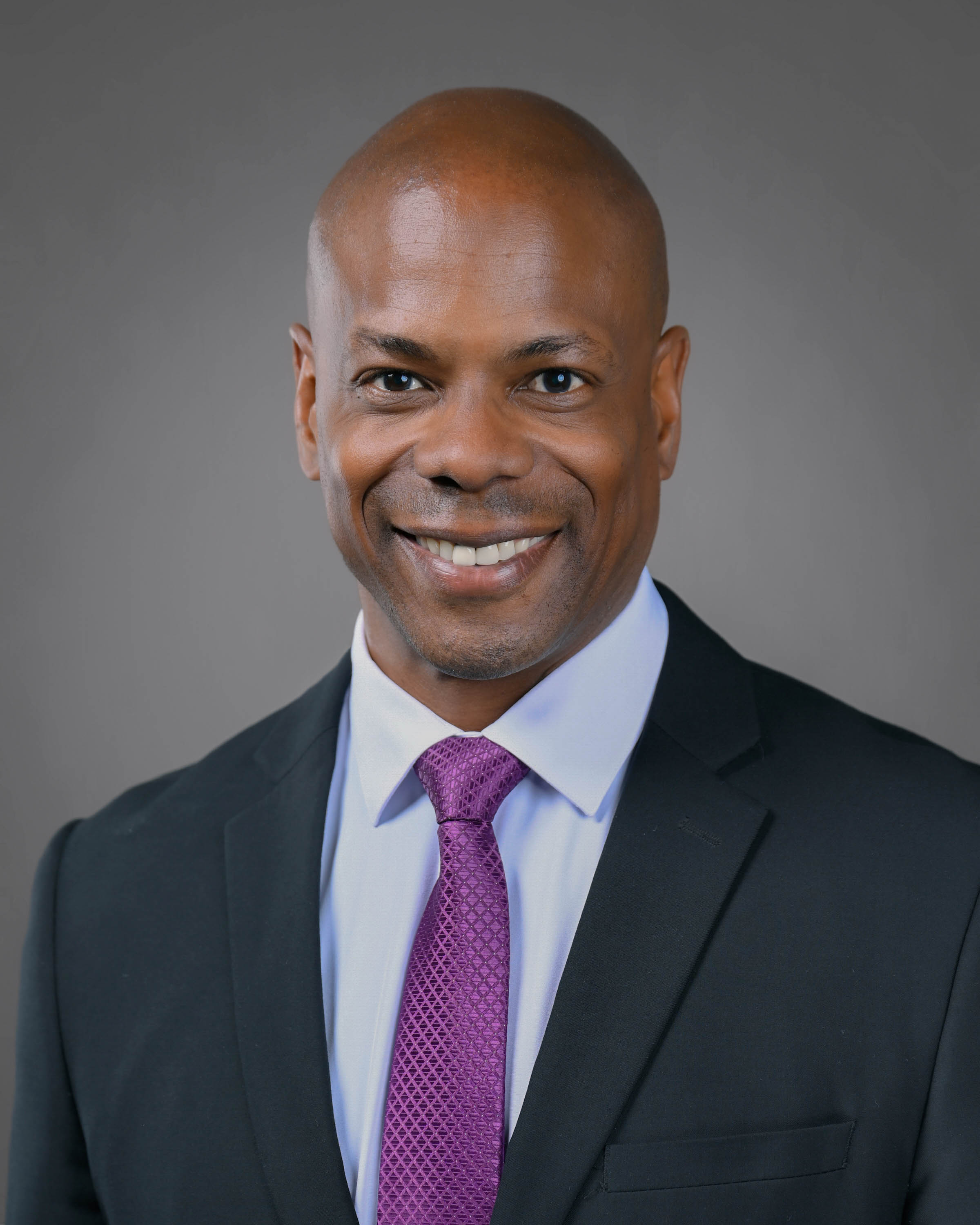 Kind looking man smiling with a dark jacket on and purple tie